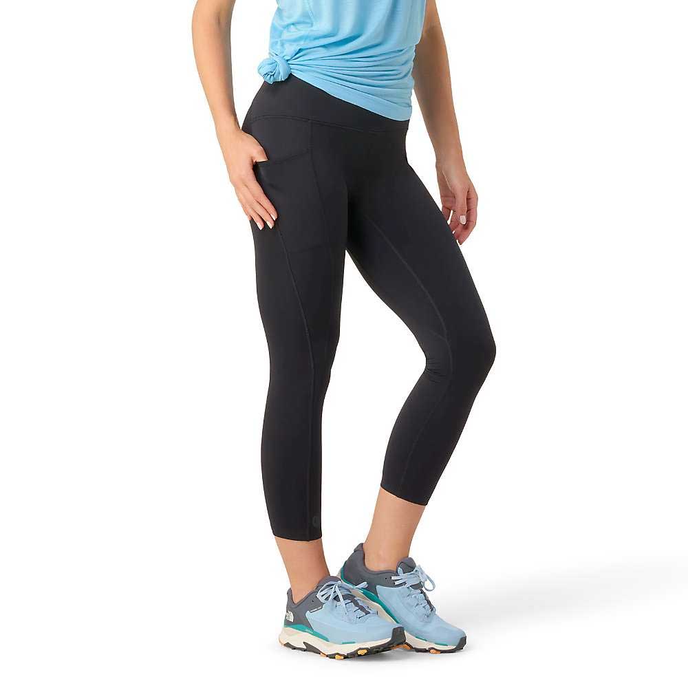 A person stands in a blue shirt, Smartwool Women’s Active 7/8 Legging, and blue athletic shoes. The torso to feet are visible, with one hand on the hip and the other at their side.
