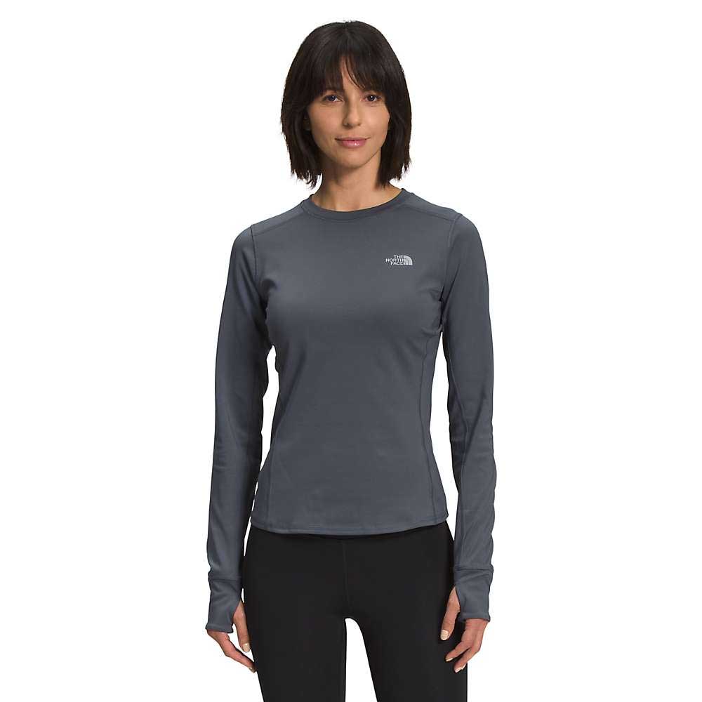 A woman wearing The North Face Womens Winter Warm Essential Crew long-sleeve shirt in dark gray and black leggings stands facing forward against a white background, carrying a sleek 30-liter backpack.