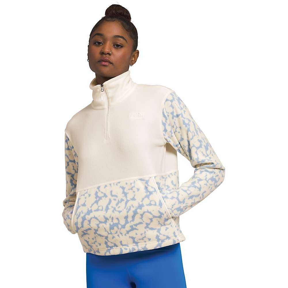 A woman with her hair in a bun wears The North Face Womens Alpine Polartec 100 1/4 Zip Cowl Top in cream and light blue, standing with hands in the front pocket and pairing the cozy pullover with bright blue leggings.