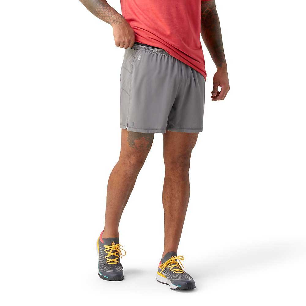 A person in a red shirt, Smartwool Mens Active Lined 5 Inch Short, and gray running shoes with yellow laces stands against a white background, lifting their shirt hem slightly.