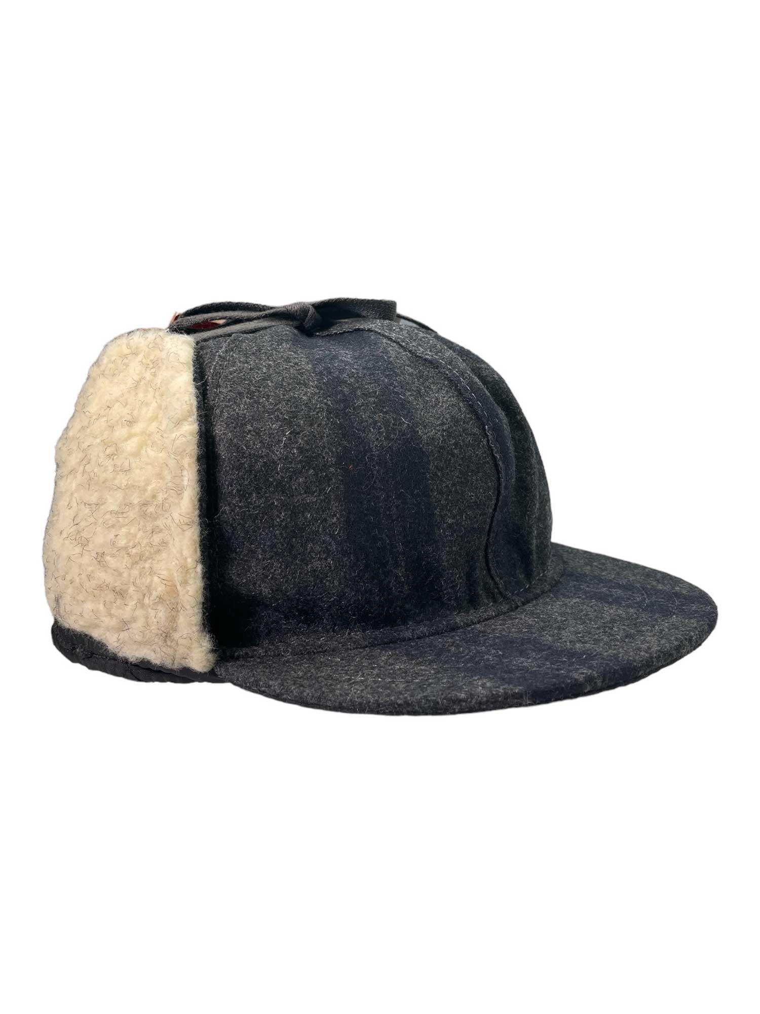 The Stormy Kromer The Snowdrift Cap is a dark gray wool hat with a wide brim and a cream shearling panel covering one side and the back, shown against a plain white background.