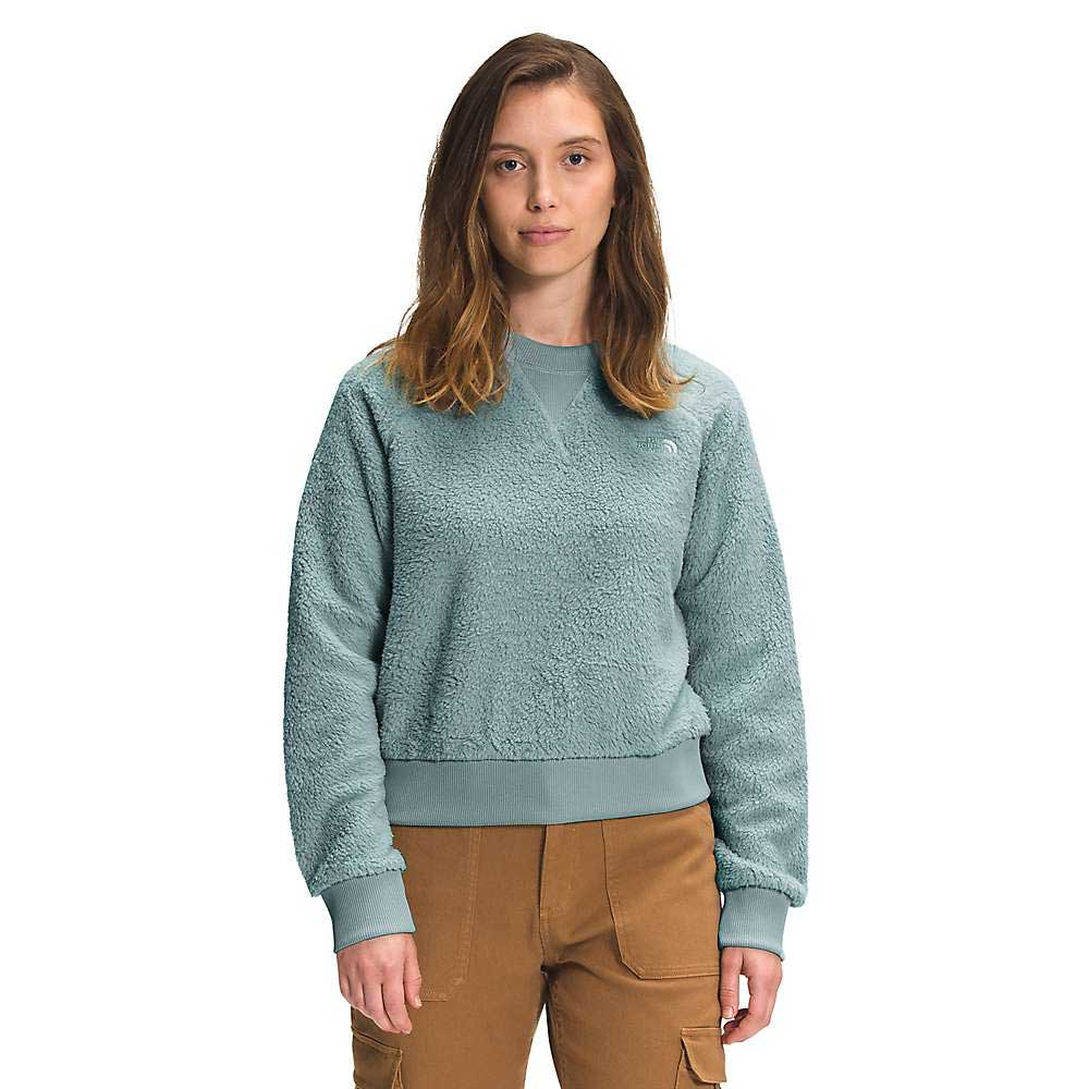 A woman with shoulder-length brown hair wears The North Face Womens Dunraven Crew Top in light teal fleece and brown pants, standing facing forward against a white background.
