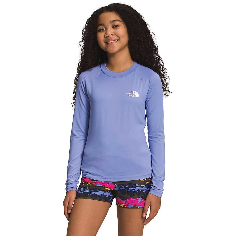A smiling girl with long curly hair wears The North Face Girls Amphibious LS Sun Tee and colorful patterned shorts, standing against a white background.