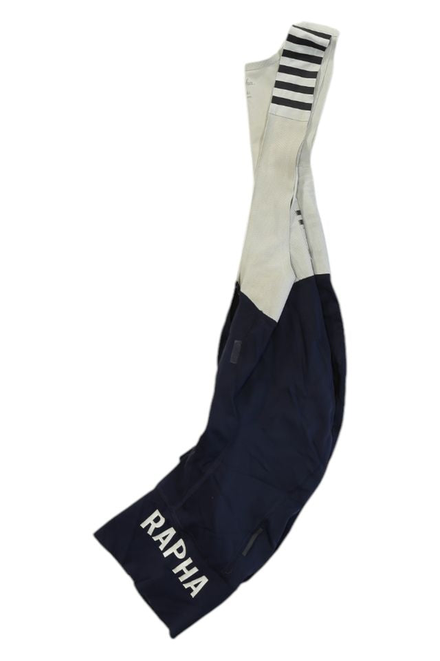 Rapha Mens Pro Team Bib Shorts II - Regular in navy blue and white with RAPHA printed in white on the leg, race-grade chamois, and black horizontal stripes on one shoulder strap, laid flat on a white background.