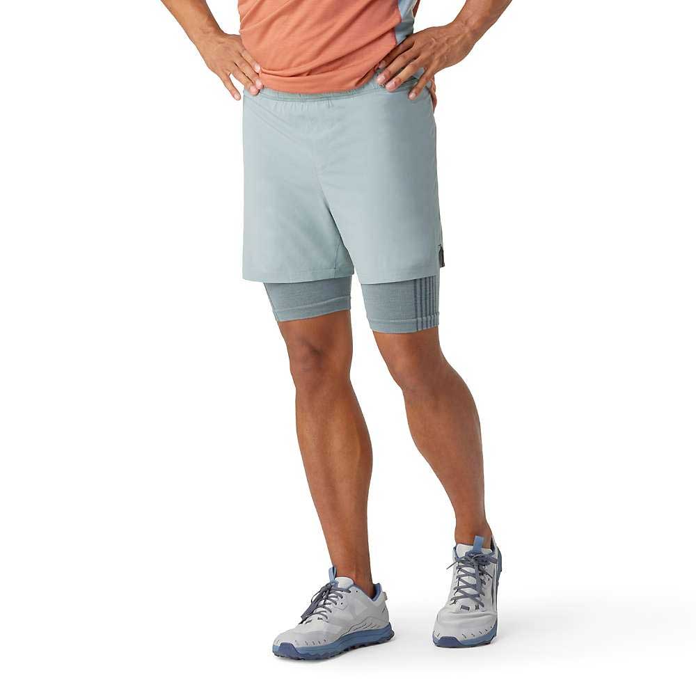 A person in a peach athletic shirt, Smartwool Men's Intraknit Active Lined 5 Inch Shorts in light gray, and light gray athletic shoes stands hands on hips against a white background.