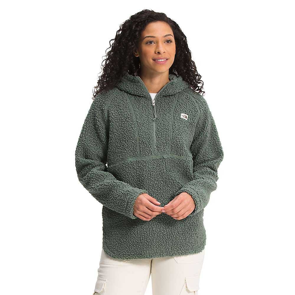 A woman with curly hair wears the Women's Ridge Fleece Tunic, a textured green pullover with a half-zip and front pocket. She stands against a plain white background, facing forward with her hands clasped.