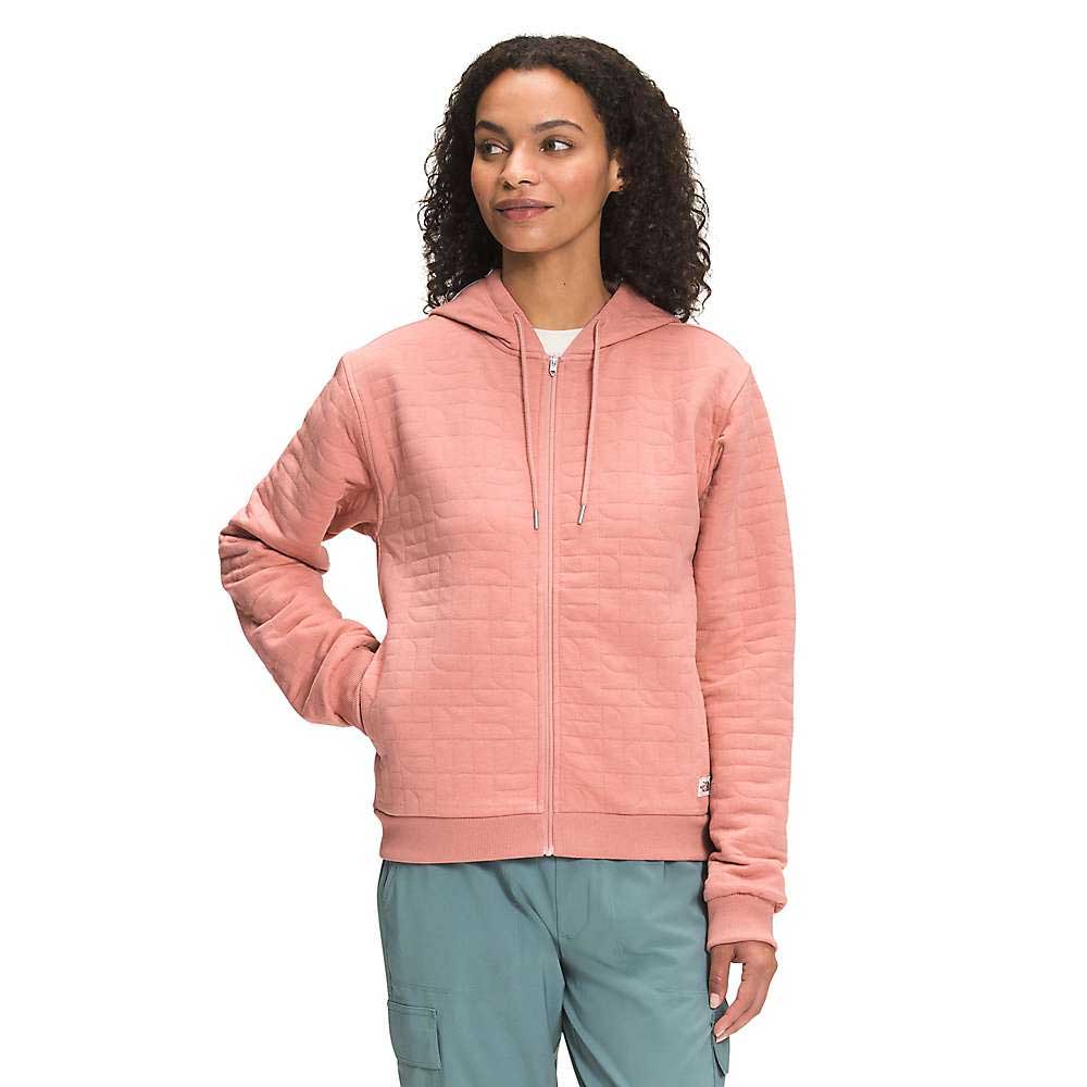 A woman stands with one hand in her pocket, smiling softly and looking to the side, dressed in The North Face Womens Longs Peak Quilted Full Zip Hoodie in light pink and light blue-green pants.