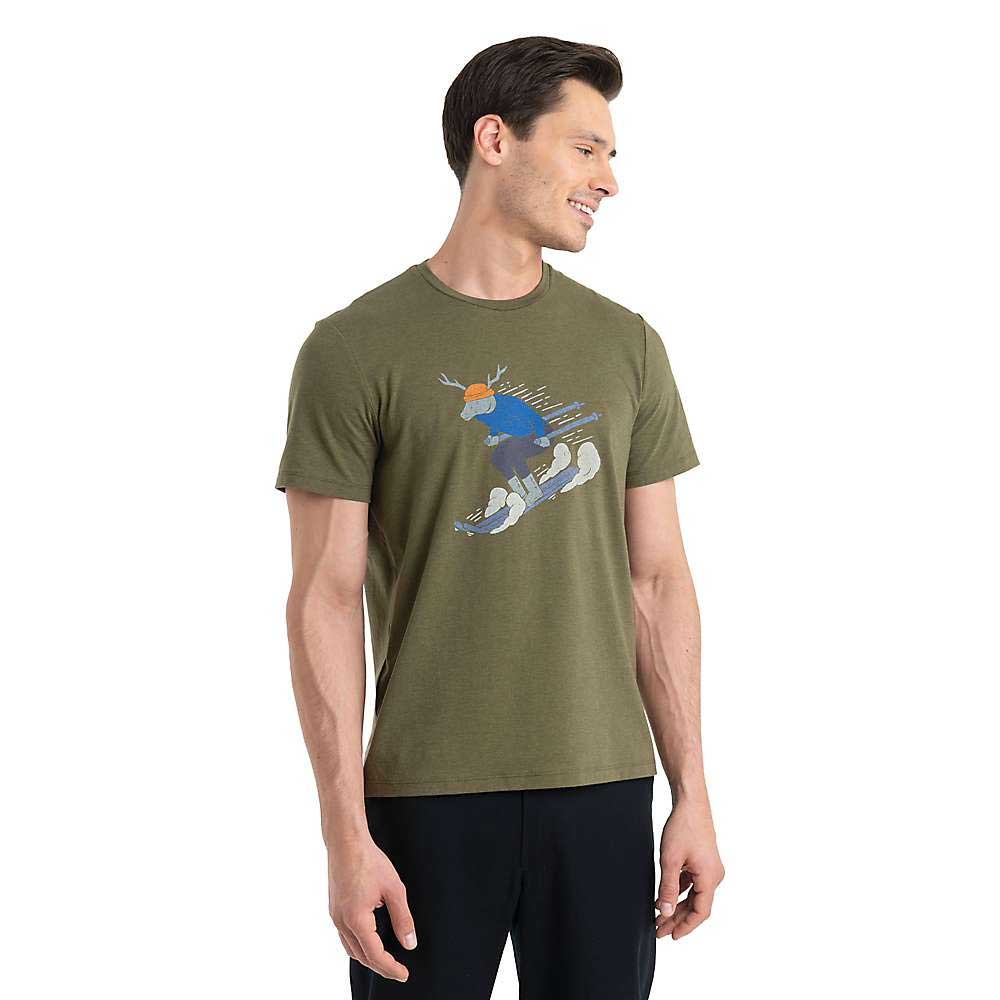 A man wears the Icebreaker Mens Merino Central Classic Ski Rider SS Tee, featuring a skiing moose graphic in olive green, paired with dark pants—ideal for hiking or casual outings—standing against a plain white background and smiling.