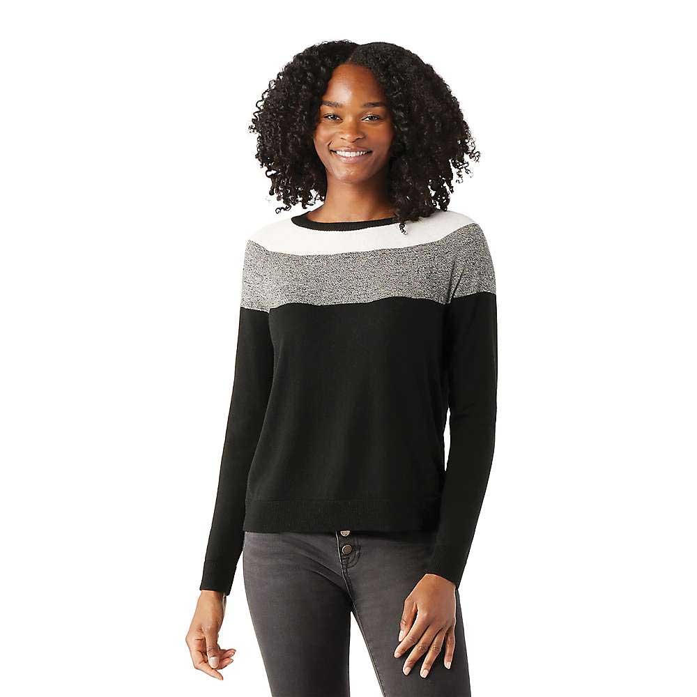 A woman with curly hair smiles while wearing the Smartwool Womens Edgewood Colorblock Crew Sweater in black, gray, and white. She stands facing forward against a plain white background with one arm relaxed at her side.