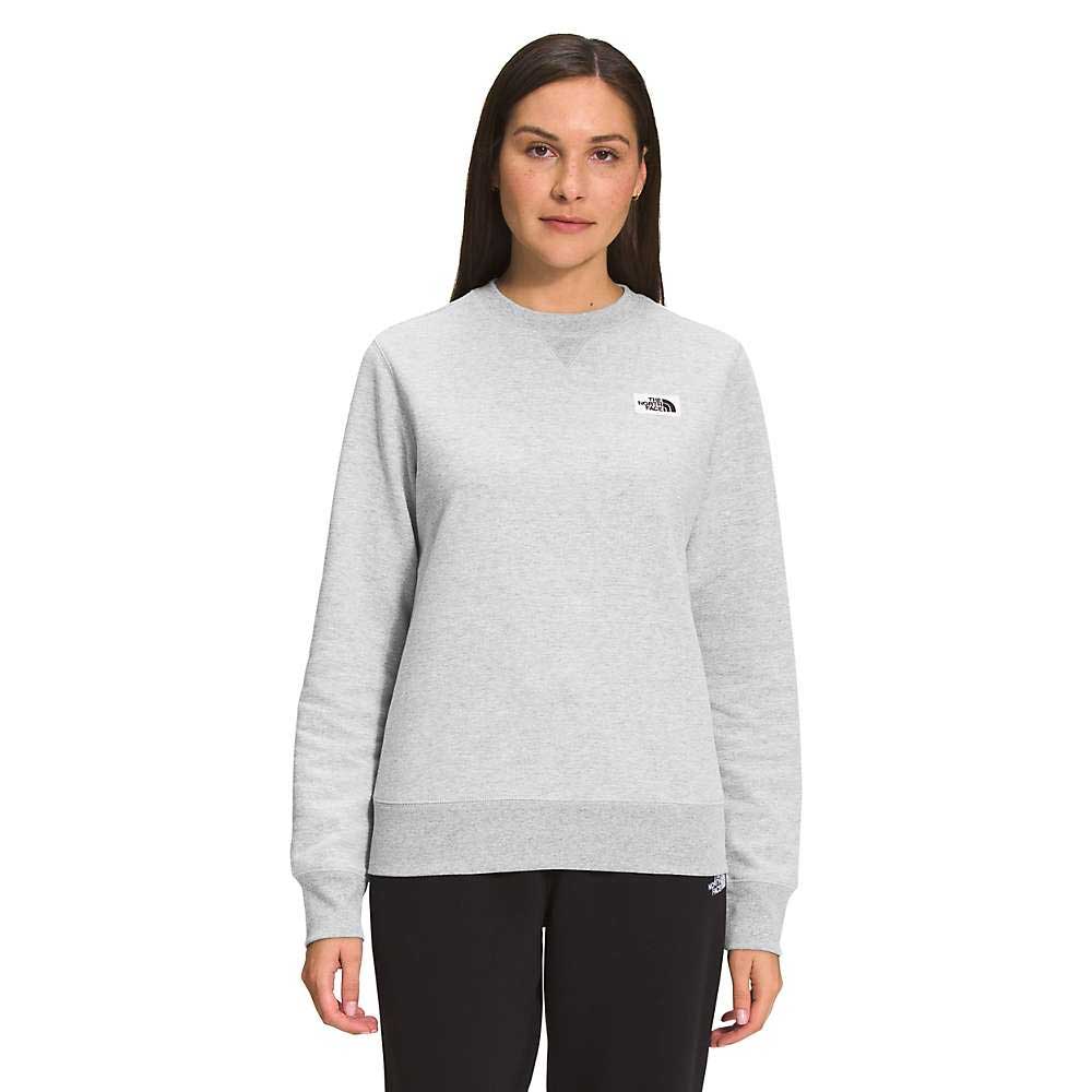 A woman faces forward in The North Face Womens Heritage Patch Crew, a light gray sweatshirt made from recycled fabric with a small left chest logo, paired with black pants against a plain white background.