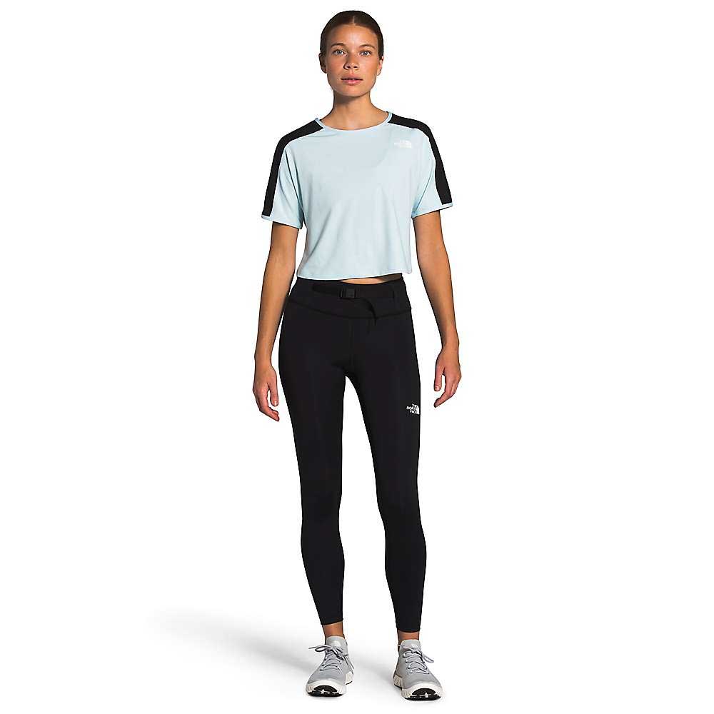 A woman faces forward, wearing a light blue short-sleeve athletic top with black accents, The North Face Womens Active Trail High-Rise Waist Pack Tight pants, and gray athletic shoes against a plain white background.