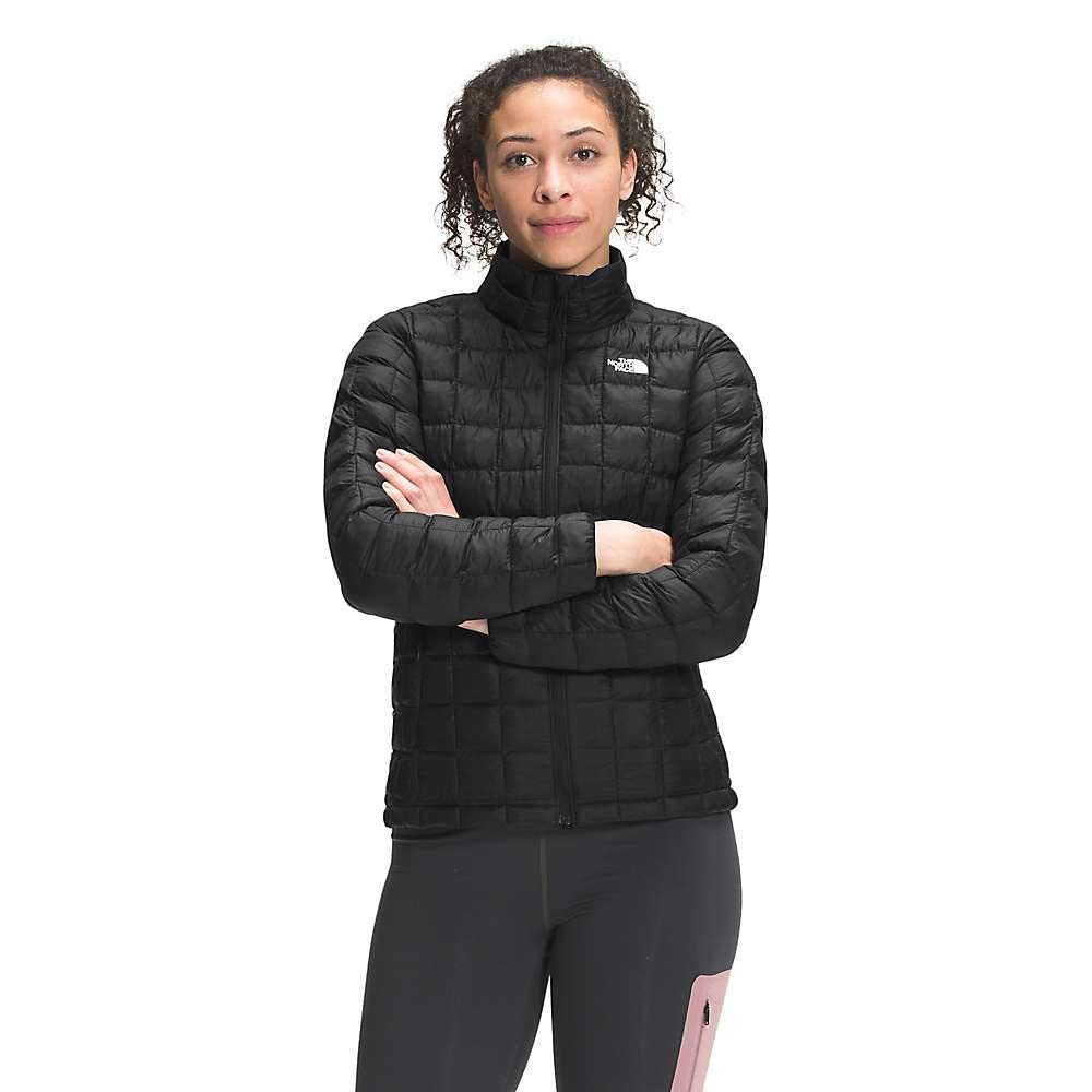 A woman stands facing the camera, arms crossed, wearing The North Face Women's ThermoBall Eco Jacket with a high collar and zip front. Made from recycled nylon with synthetic insulation, it features a small white chest logo.