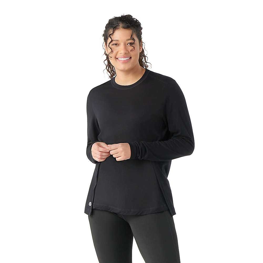 A smiling woman with curly hair stands against a white background, wearing the Smartwool Womens Active LS Crew Top in black paired with black leggings.