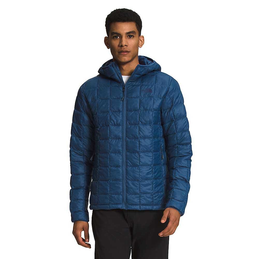 A person wears The North Face Mens ThermoBall Eco Hoodie in blue, with a quilted design and front zipper, standing against a plain white background.