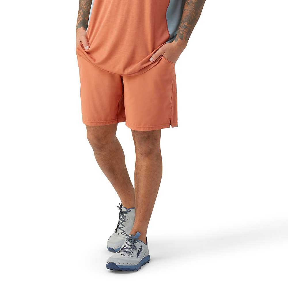 A person wearing an orange athletic shirt and Smartwool Men's Active Lined 8 Inch Shorts with hands in pockets, paired with light blue-gray hiking shoes, stands against a plain white background. The quick drying fabric is perfect for travel. Head not visible.