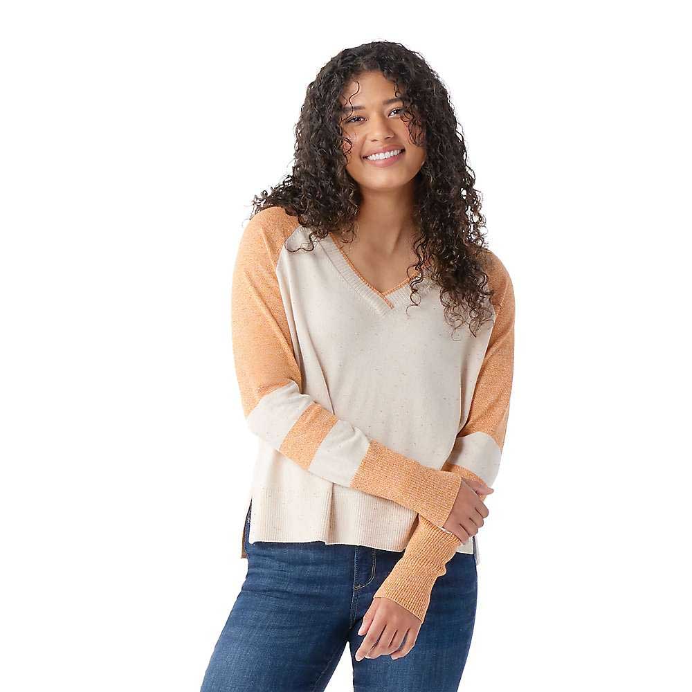 Woman with long curly hair smiles, standing against a white background in blue jeans and the beige and orange Smartwool Womens Edgewood V-Neck Sweater, her arms relaxed in front of her.