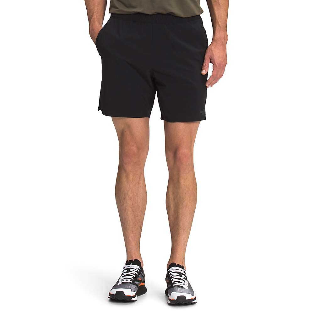 A person in a green shirt, The North Face Mens Wander 7 Inch Short (black), and black-and-white running shoes stands with hands in pockets against a plain white background.