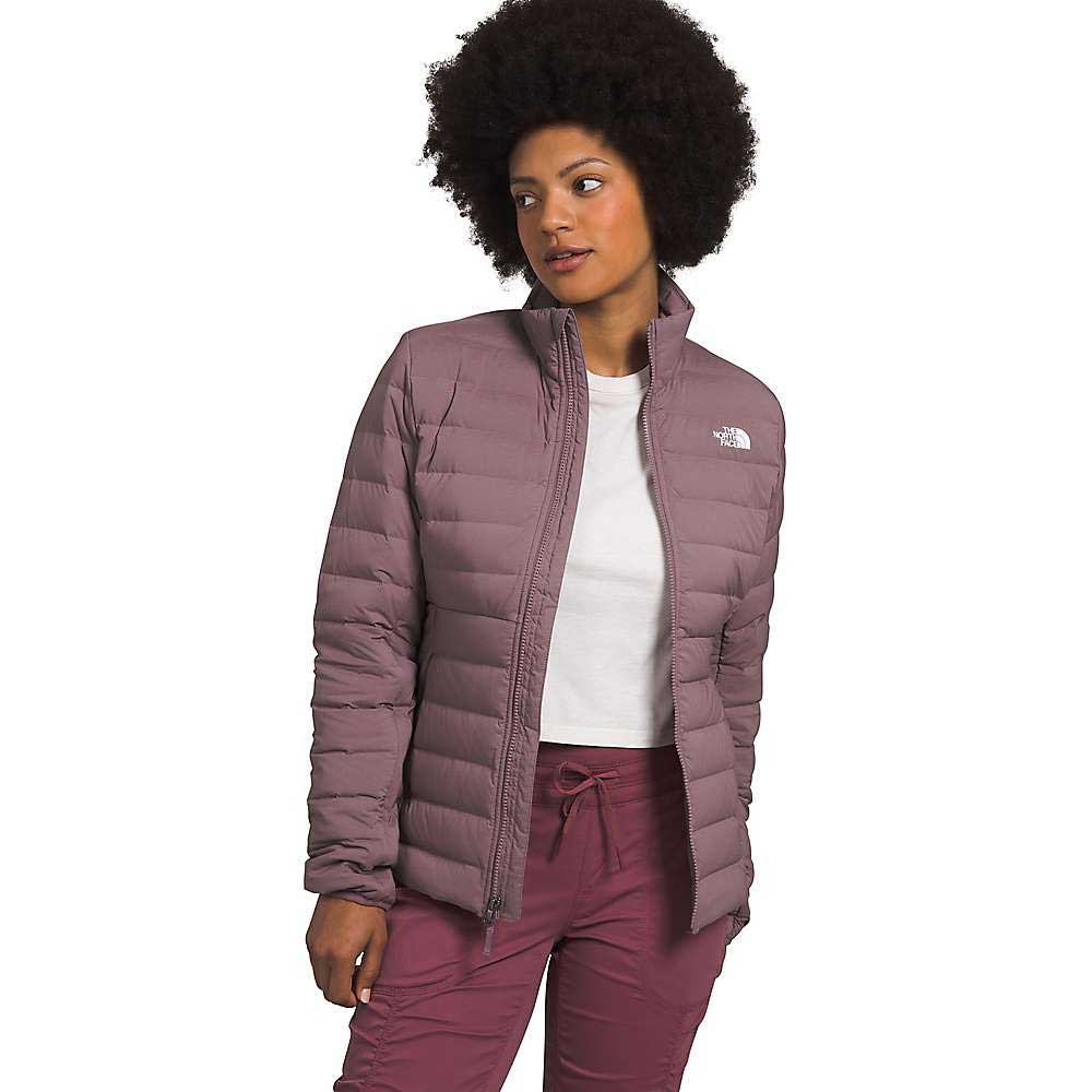 A woman with curly hair wears The North Face Women's Belleview Stretch Down Jacket in mauve, made with recycled waterfowl down, over a white t-shirt and matching mauve pants. She stands with one hand in her pocket against a plain white background.