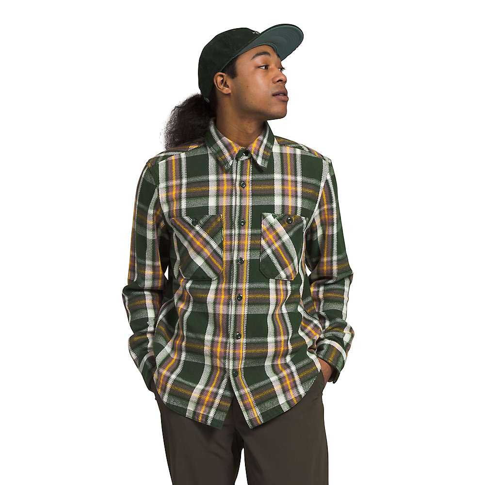 A person wearing The North Face Mens Valley Twill Flannel Shirt in green plaid, with brown pants and a green cap, stands hands-in-pockets, looking to the side against a plain white background.