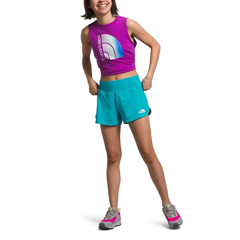 A person stands smiling with hands on hips against a white background, wearing a sleeveless purple crop top, The North Face Girls Amphibious Knit Short in turquoise, and gray and pink sneakers.