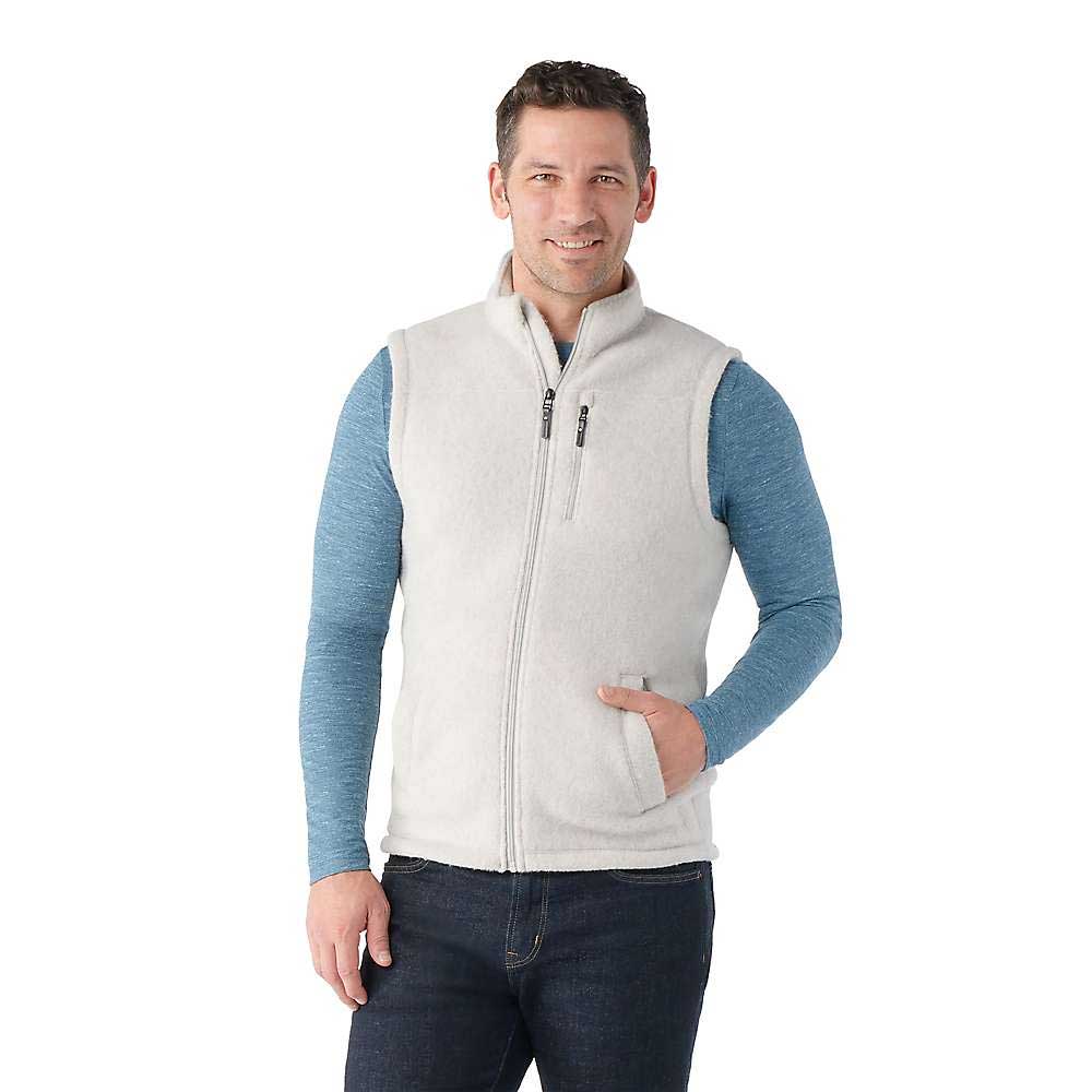 A man in a Smartwool Men's Hudson Trail Fleece Vest, layered over a blue long-sleeve shirt and dark jeans, smiles with one hand in his vest pocket against a plain white background.