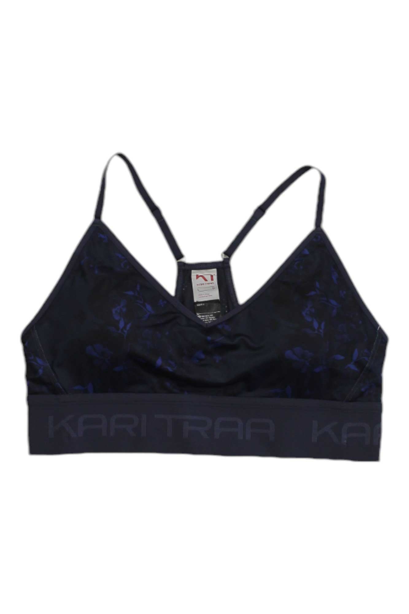 The Kari Traa Womens Var Printed Sports Bra is black with thin adjustable straps, a subtle dark blue floral pattern, a wide elastic KARI TRAA band, and a light support fit. The inside has a visible garment tag.