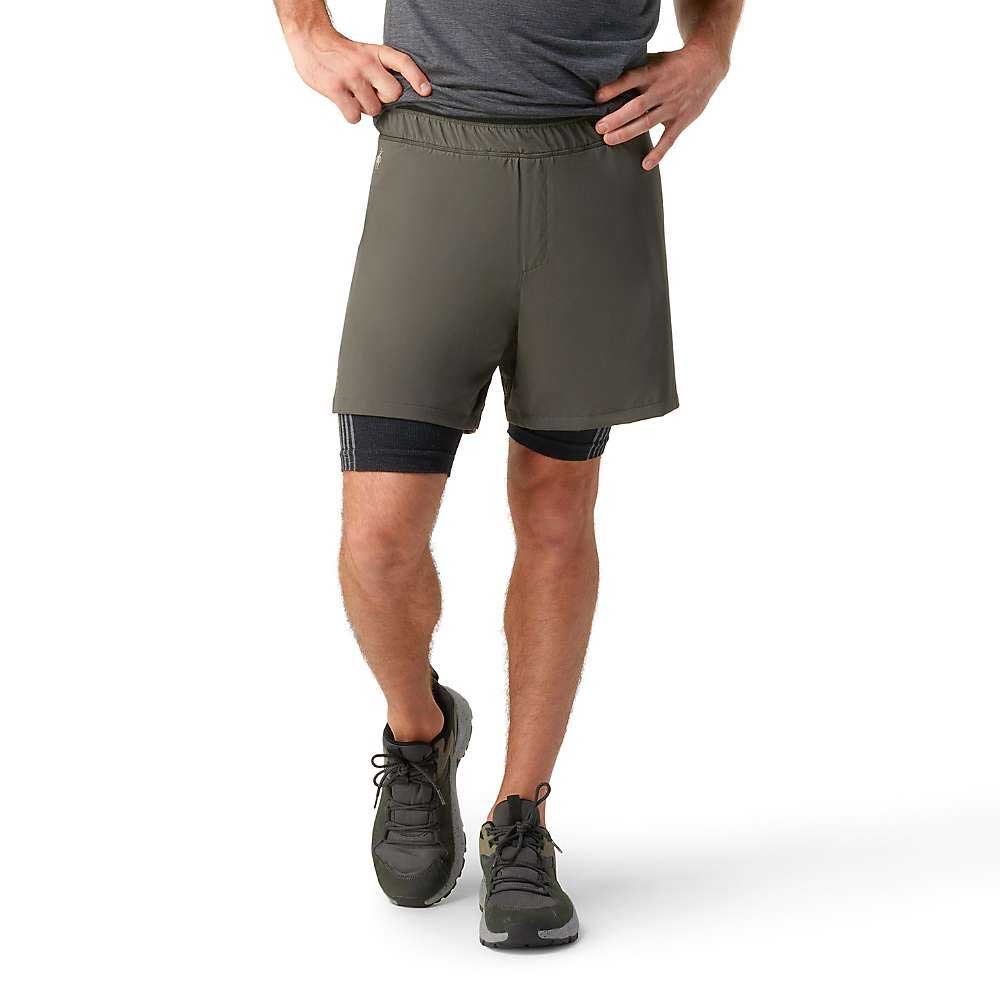 Wearing a grey tee, Smartwool Mens Intraknit Active Lined 5 Inch Shorts in olive over black compression shorts, and grey running shoes, a person stands with one hand on their hip and the other resting on their thigh.