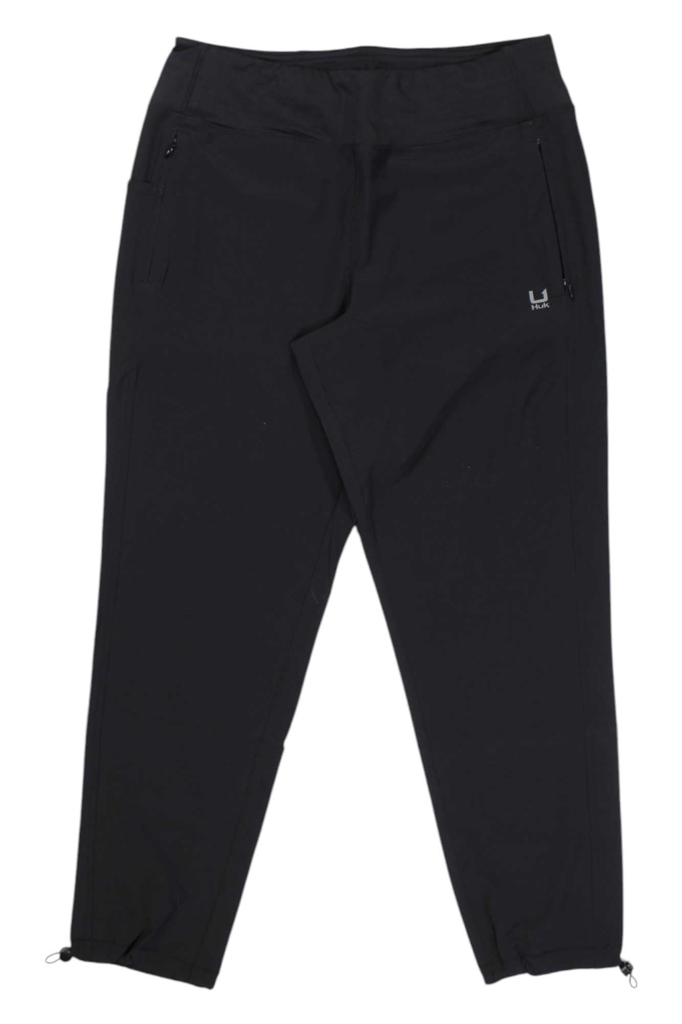 The Huk Womens Cedros Pant in black features zippered side pockets, adjustable drawstring ankle cuffs, and a small logo on the left hip. Designed for outdoor adventures, these versatile pants are perfect for hiking or any active outing. Shown laid flat on a white background.
