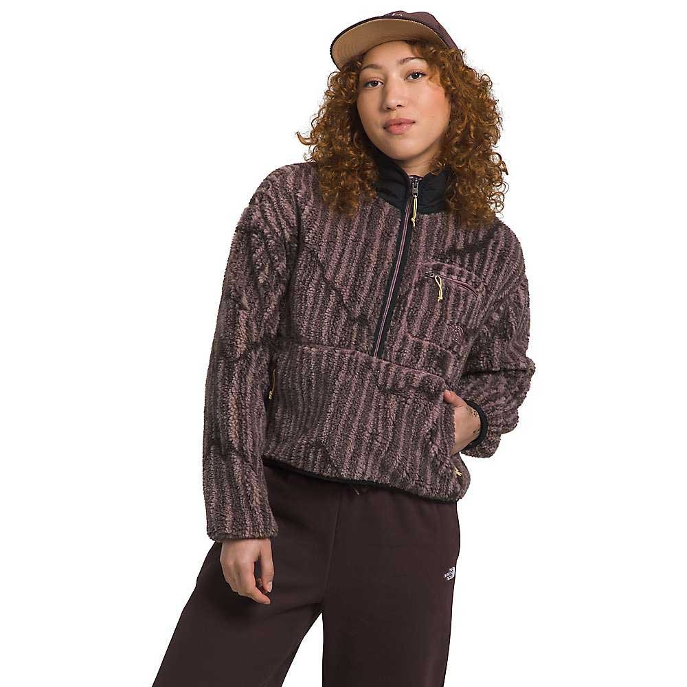 A person with curly hair wears The North Face Women's Extreme Pile Pullover, styled with dark pants and a two-tone baseball cap, while standing against a white background.