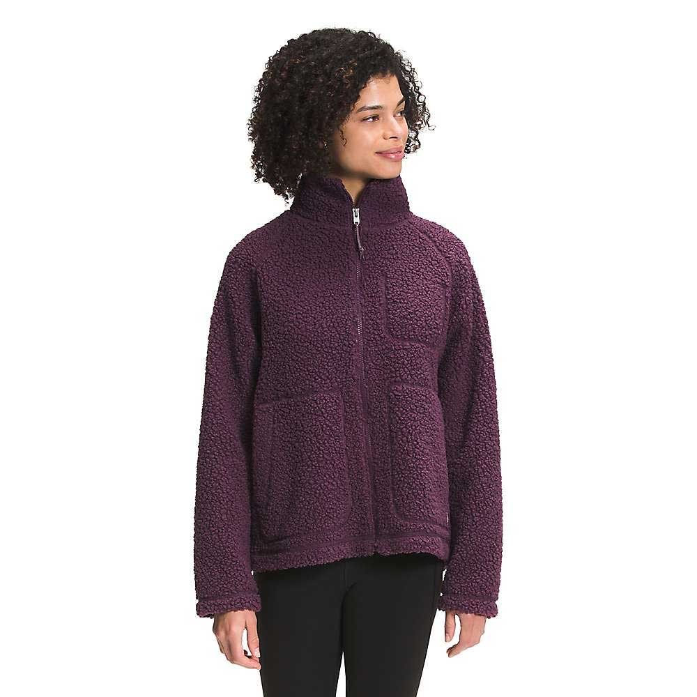 A woman with curly hair wears The North Face Womens Ridge Fleece Full Zip Jacket in dark purple, featuring large front pockets. She stands facing left, smiling slightly, against a plain white background.