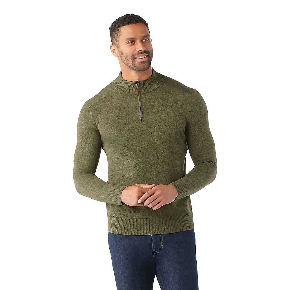 A man in a fitted, olive green Smartwool Men's Sparwood Half Zip Sweater and dark blue jeans stands smiling with his hands loosely clasped before him against a plain white background.