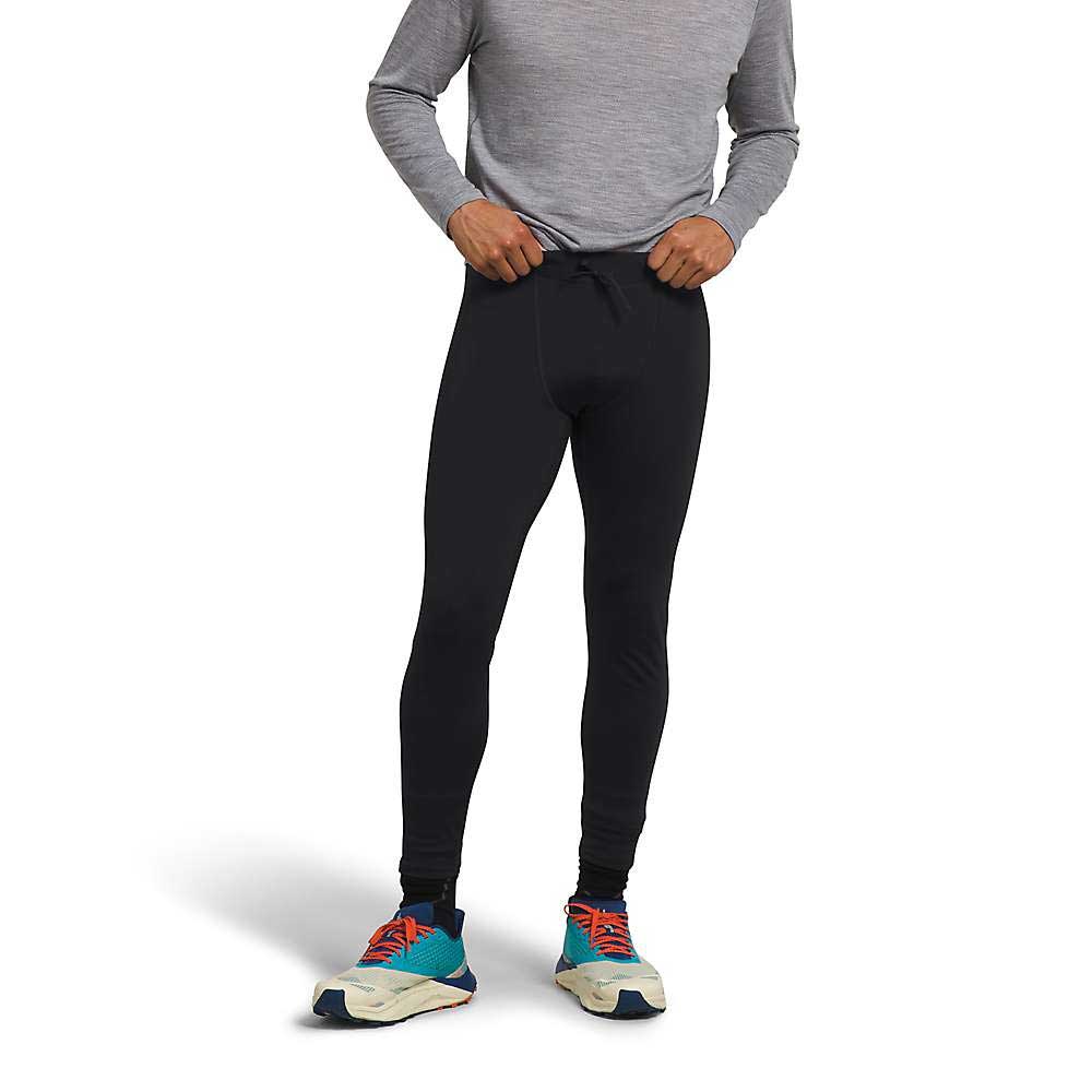 A person wearing The North Face Mens Winter Warm Essential Legging in black and a gray long-sleeve shirt stands against a white background, adjusting the waistband of the leggings.