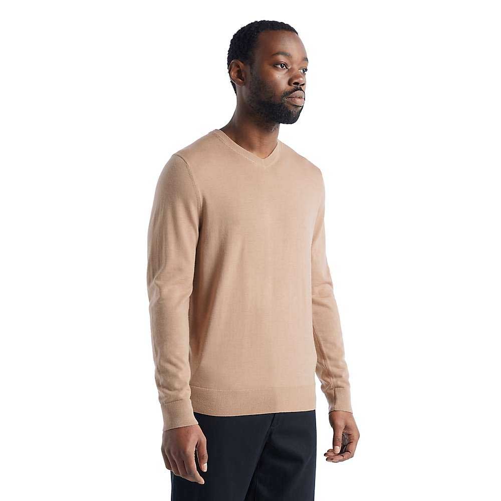 A man stands against a plain white background, wearing the Icebreaker Men's Wilcox LS V Neck Sweater in beige and black pants, facing slightly to the side with a neutral expression.