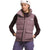 A person wears The North Face Women's Gotham Vest in light purple over a dark long-sleeve shirt, paired with purple pants and a white The North Face logo beanie, standing hands-in-pockets against a plain white background.