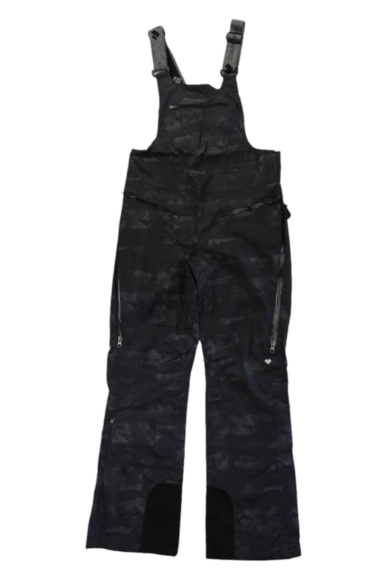 The Obermeyer Womens Bliss Bib Pant features a black and dark gray camo pattern, adjustable shoulder straps, a zippered chest pocket, side leg zippers, insulation for warmth, and reinforced black inner ankles for durability.