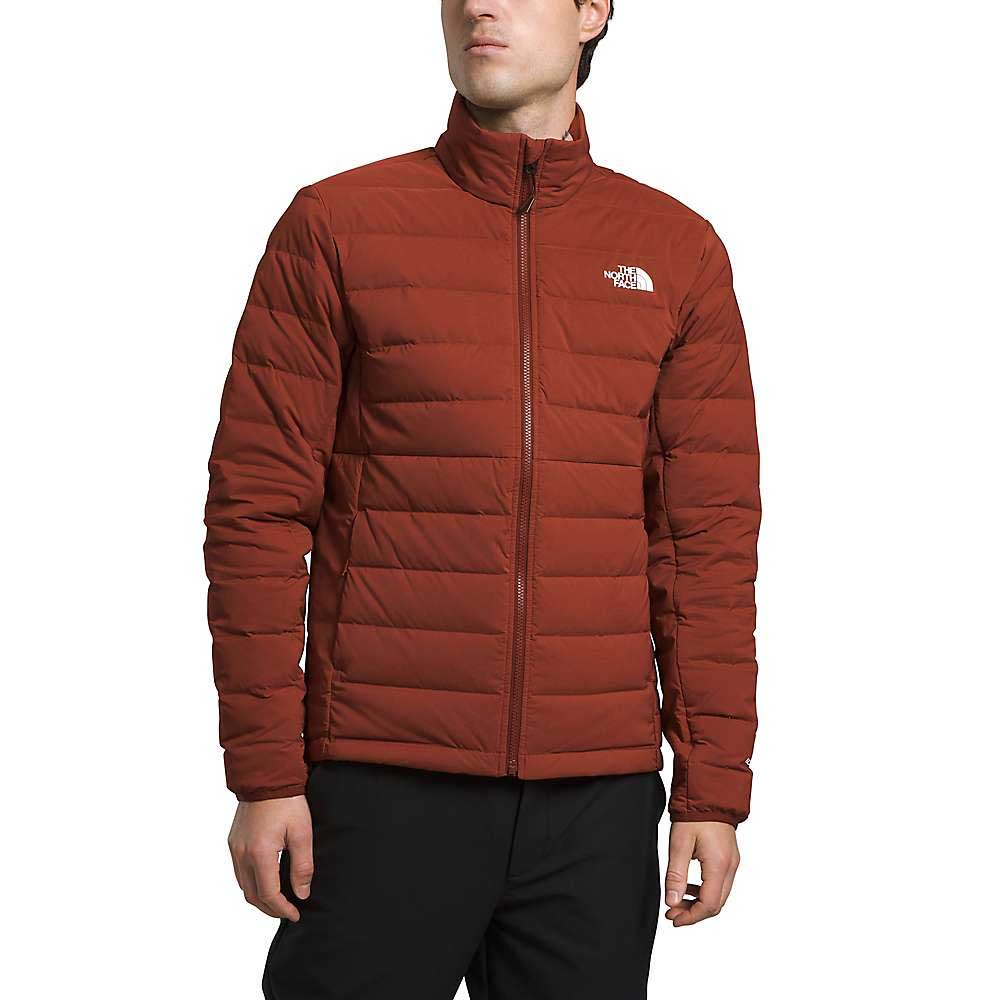 A person stands against a plain white background wearing The North Face Mens Belleview Stretch Down Jacket in rust, featuring recycled down insulation and a front zipper, paired with black pants.