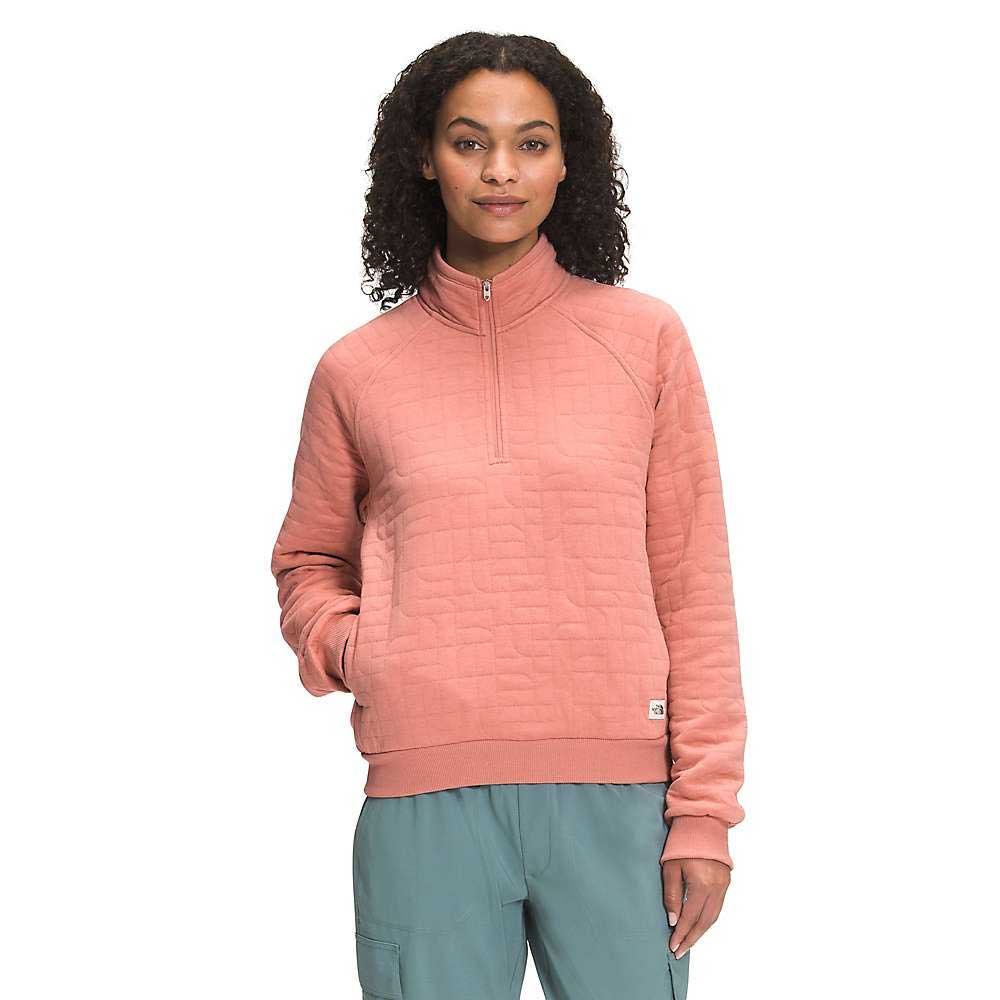 A woman with curly hair wears The North Face Women's Longs Peak Quilted 1/4 Zip Jacket in pink and teal pants, standing with one hand in her pocket and smiling slightly at the camera against a plain white background.