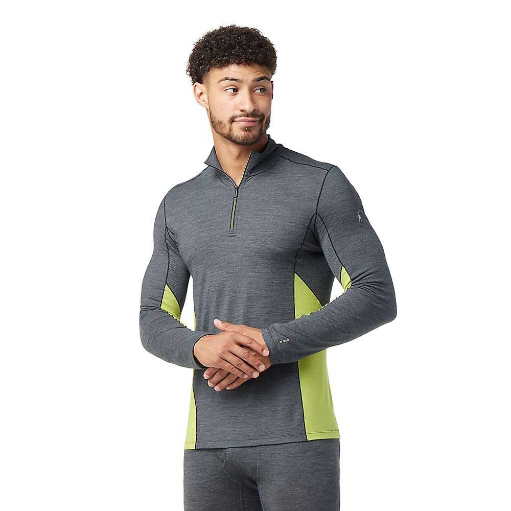 A man wearing the Smartwool Men’s Merino Sport 150 LS 1/4 Zip Top in gray with yellow-green side panels stands with hands clasped, looking aside. The athletic top features a front zipper and is paired with matching gray athletic pants.