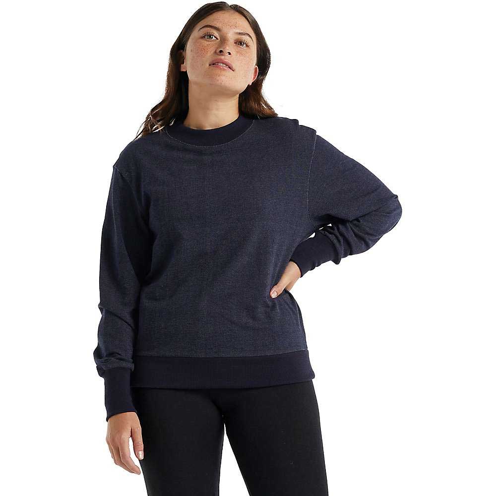 A woman with long brown hair stands confidently, wearing the Icebreaker Womens Central LS Sweatshirt in dark blue and black leggings, ready for her next travel adventure.
