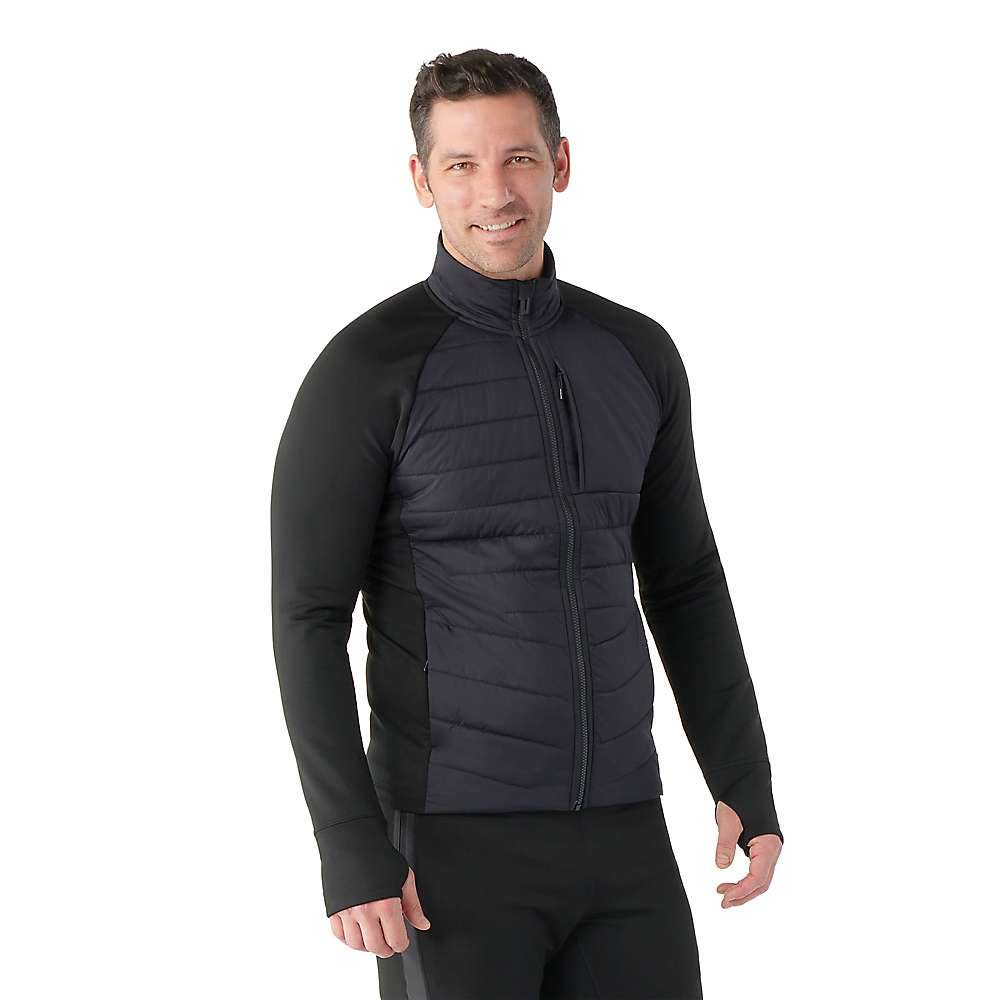A man wears a Smartwool Men's Smartloft Jacket, a fitted black insulated jacket with quilted panels and long sleeves, standing and smiling against a plain white background.