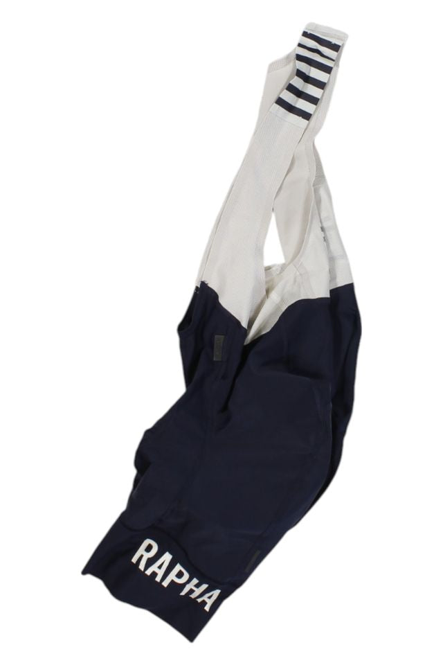 The Rapha Mens Pro Team Bib Shorts II - Regular in navy blue and white feature a race-grade chamois, RAPHA logo on one leg, and striped detailing on the shoulder strap.