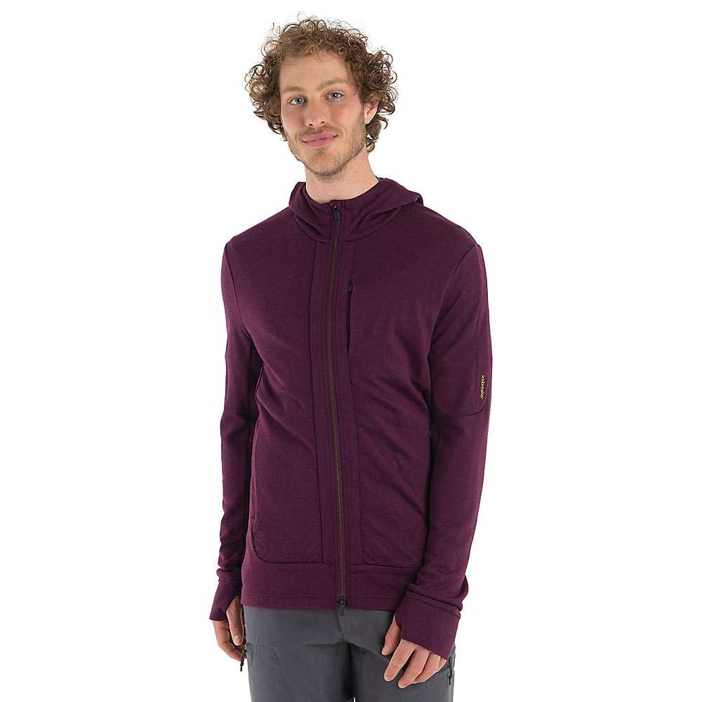 A smiling man with curly hair wears the Icebreaker Mens Quantum III LS Zip Hoodie, dark purple and long-sleeved, paired with gray pants, standing against a plain white background.