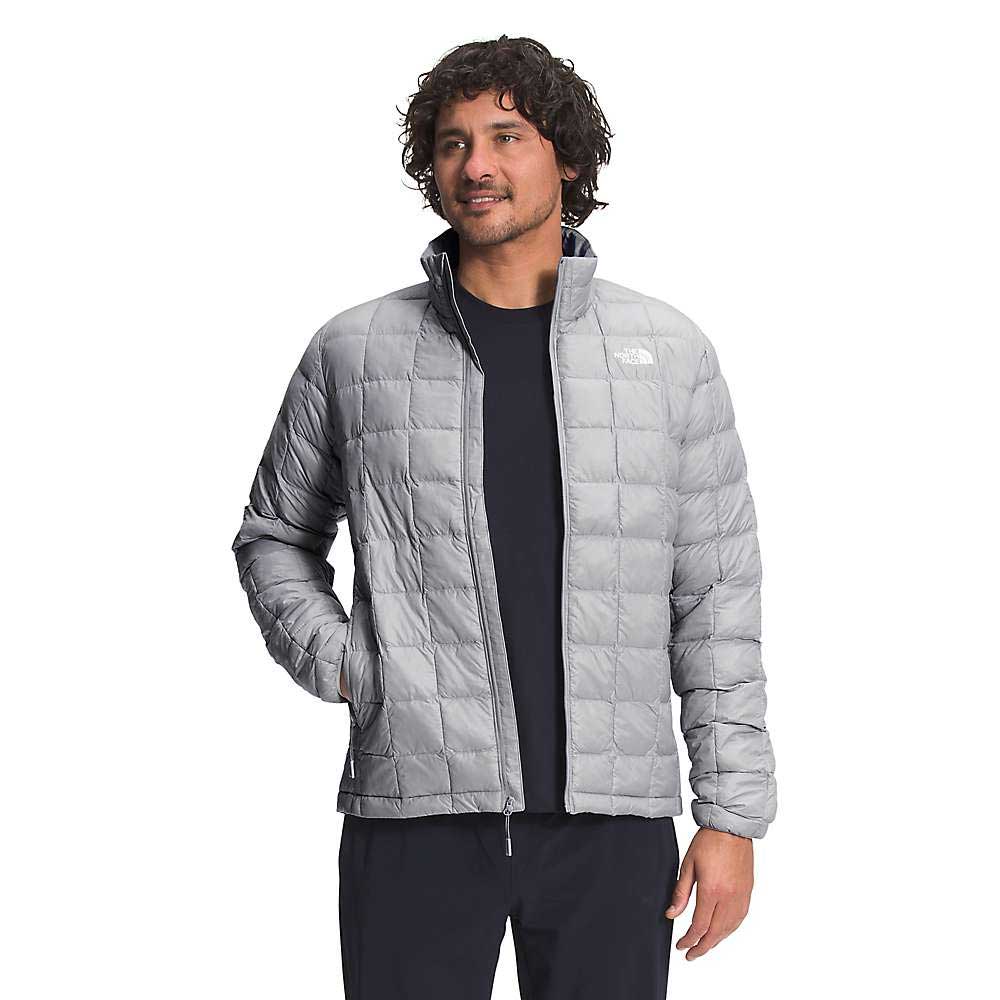 A man with curly hair wears The North Face Mens ThermoBall Eco Jacket in light gray, made from recycled nylon with a front zipper and chest logo, standing with one hand in his pocket against a white background.