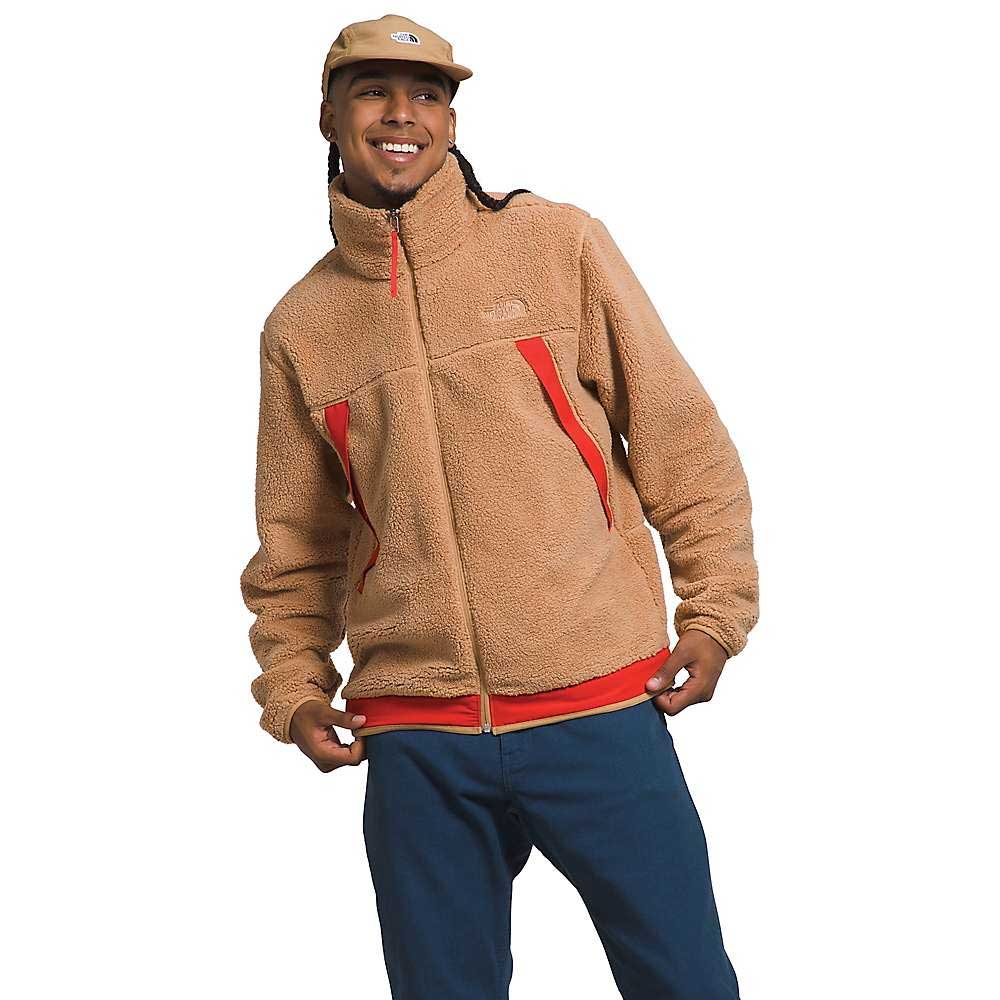 A person wears The North Face Men's Campshire Fleece Jacket in tan with red accents, paired with blue pants and a tan cap, smiling while holding the jacket’s hem against a plain white background.