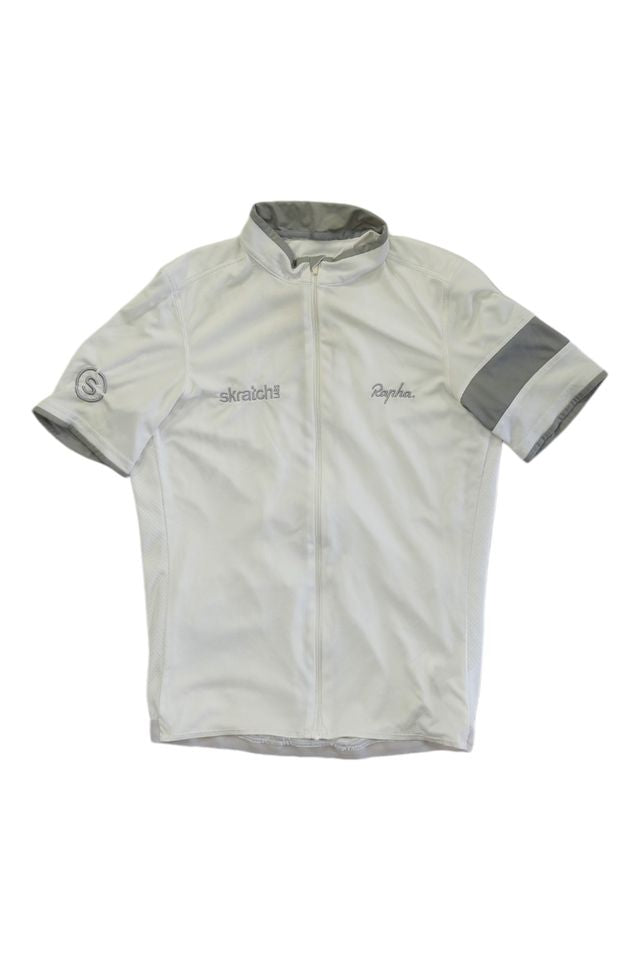 The Rapha Mens Super Lightweight Jersey is a light gray, short-sleeve cycling jersey with a full front zipper, high collar, gray left sleeve stripe, Skratch and Rapha chest logos, and a circular logo on the right sleeve.