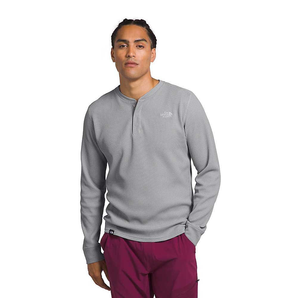 A person wears The North Face Mens Canyon Fog Thermal LS Henley—a light gray, patterned long-sleeve shirt—with maroon pants, standing casually with one hand in their pocket against a plain white background.