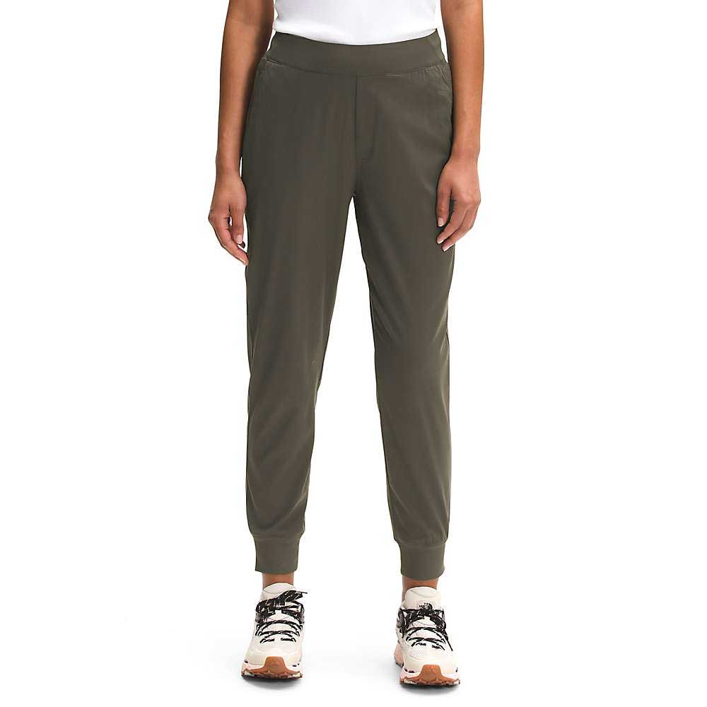 A person wears The North Face Womens Aphrodite Jogger in olive green, quick-dry fitness pants, with a white shirt and white sneakers featuring beige soles, standing against a plain white background. Image is cropped at the shoulders.