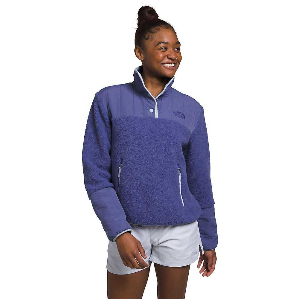 A smiling woman with her hair in a bun wears The North Face Womens Cragmont Fleece 1/4 Snap Top in blue, made from recycled polyester with a high collar and zippered pockets, paired with light grey shorts against a white background.