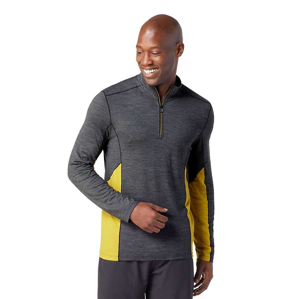 A man smiles while wearing the Smartwool Mens Merino Sport 150 LS 1/4 Zip Top—an athletic dark gray pullover with yellow accents on the sides and sleeves—as he looks down to his left.