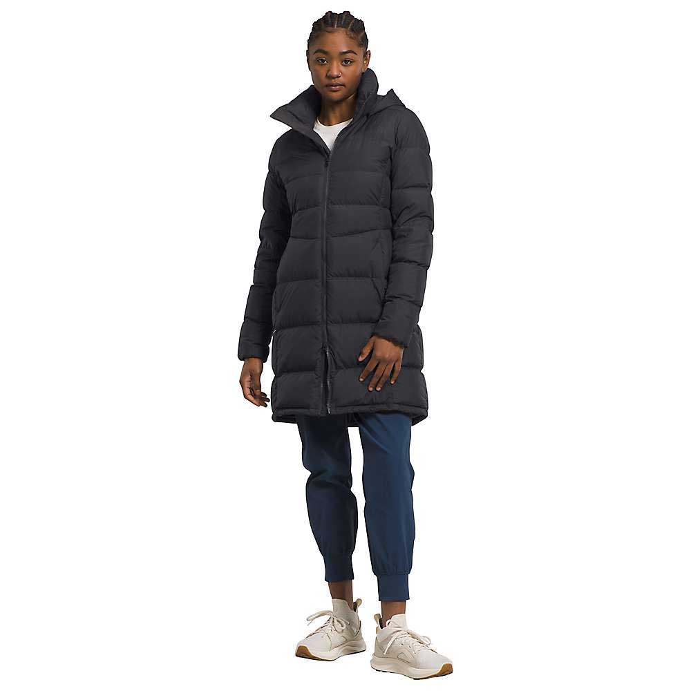A woman stands facing forward against a white background, wearing a long black The North Face Womens Metropolis Parka, blue cuffed pants, white sneakers, and a white shirt.